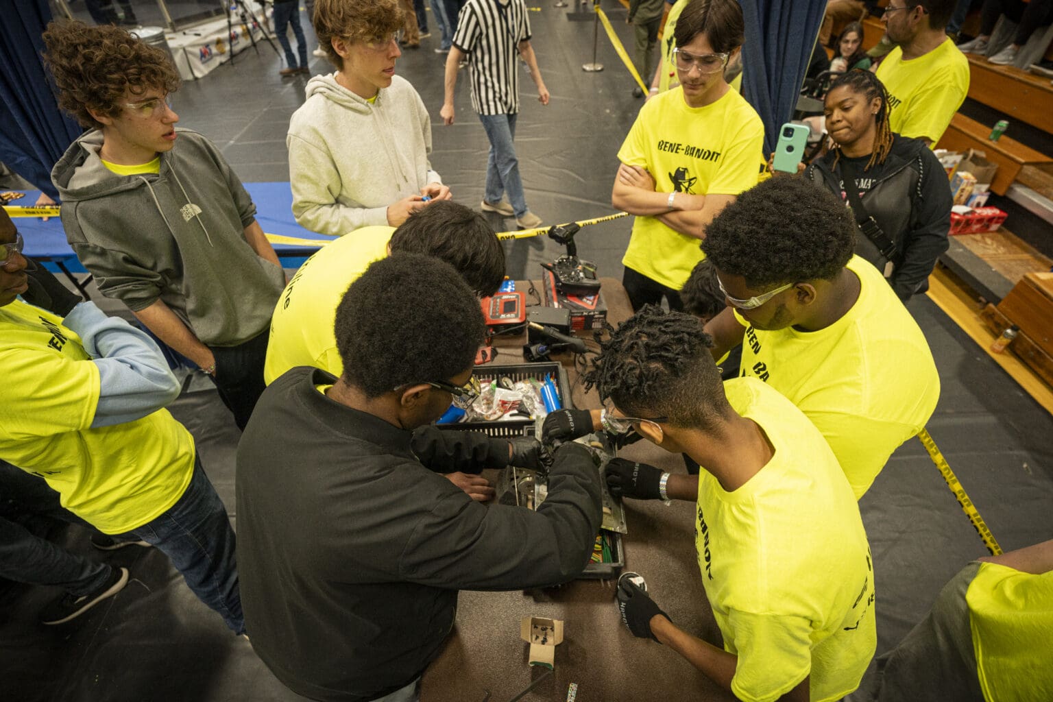 Benedictine robotics students in safety glasses and work gloves, crouched around their robot at a competition with their hands in the mechanism and a tool tray open beside them.
