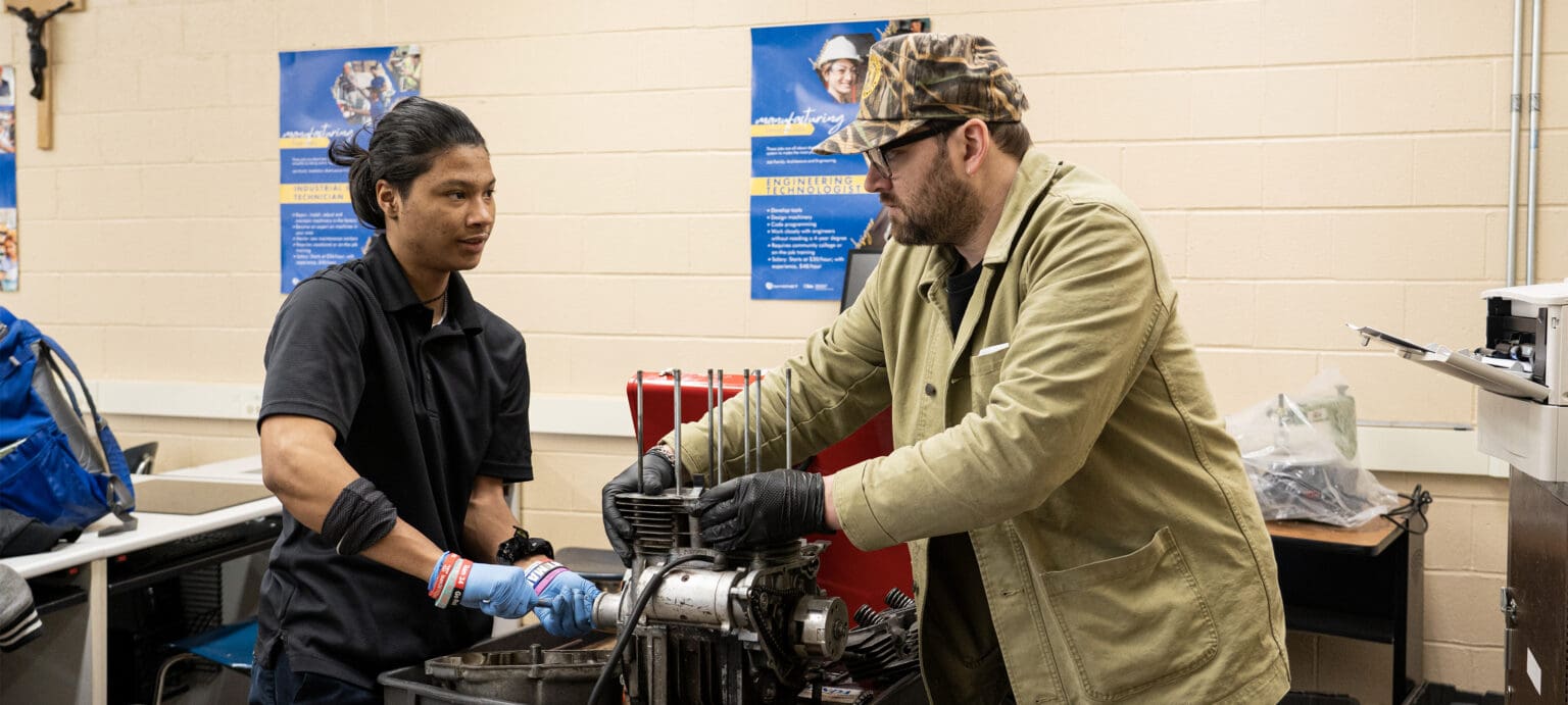 A Benedictine student in work gloves and his instructor with their hands on a stripped motorcycle engine in the school shop, manufacturing career posters on the wall behind them.