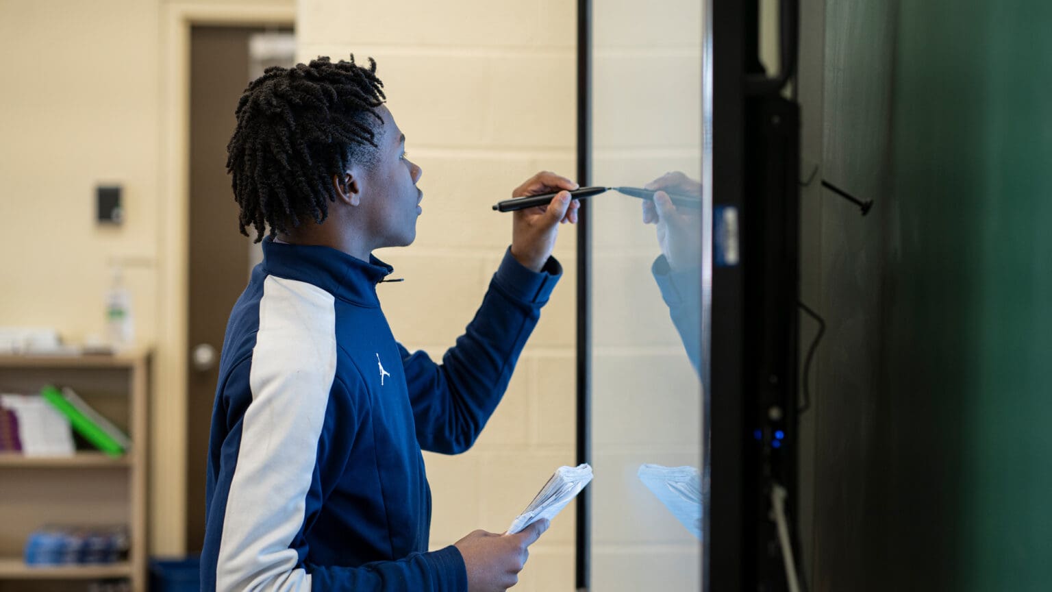 A Benedictine student at the board with a stylus in one hand and his notes in the other, working through his findings for the class.