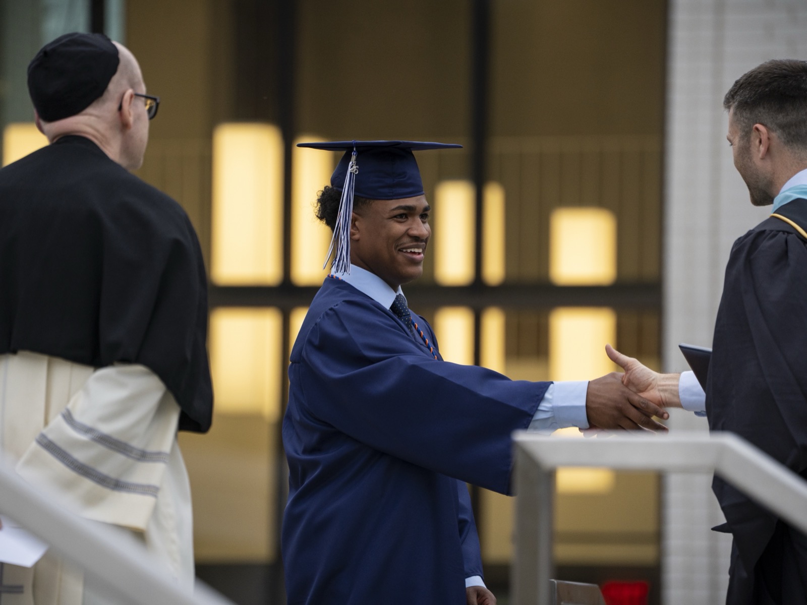 A Benedictine High School Class of 2026 graduate in a navy cap and gown shakes hands as he receives his diploma at commencement, with a Benedictine monk looking on.